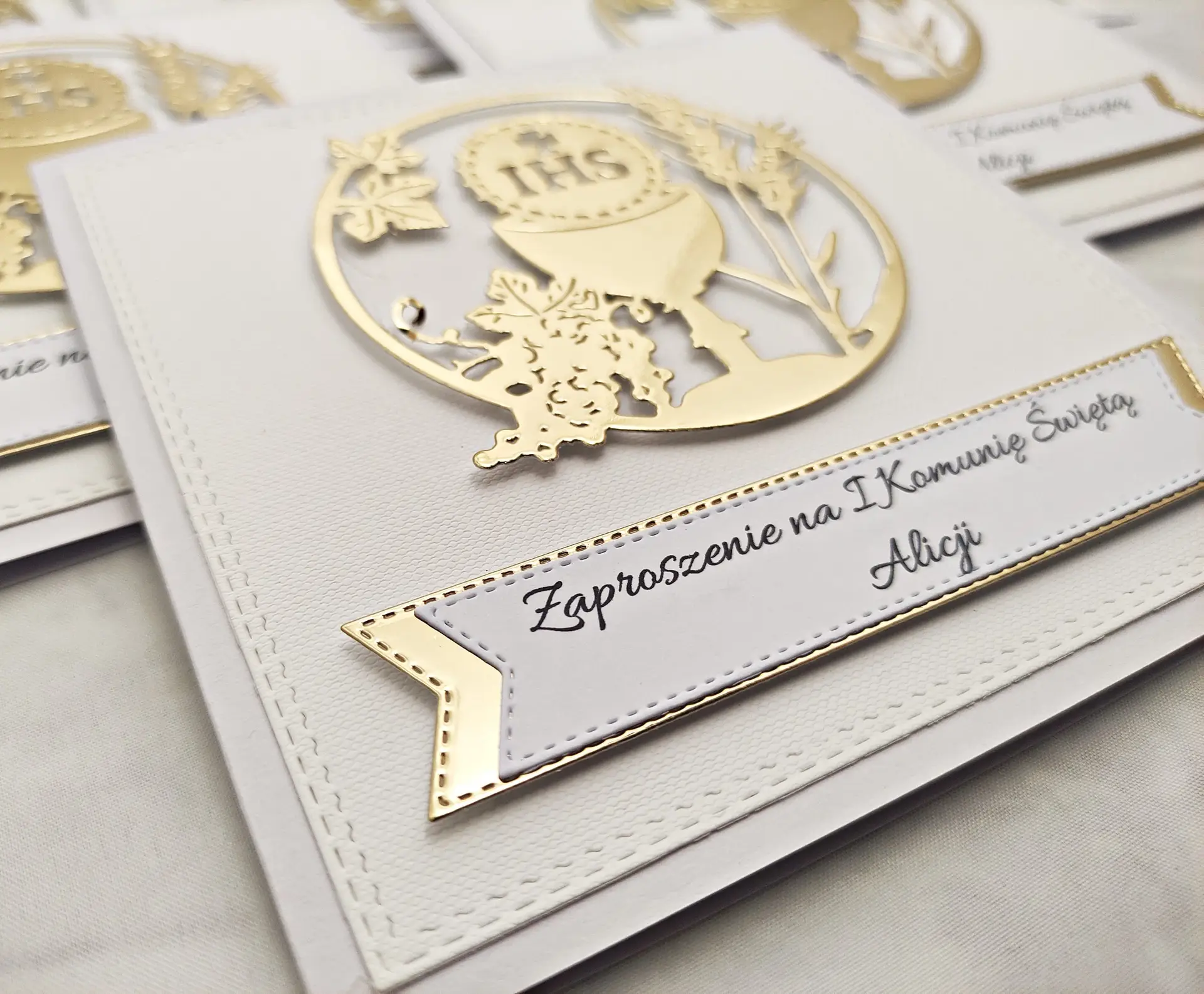 Zbliżenie detali zaproszenia komunijnego z kielichem w okręgu i napisem zaproszenie, na białym papierze wizytówkowym, subtelne złote wykończenia.