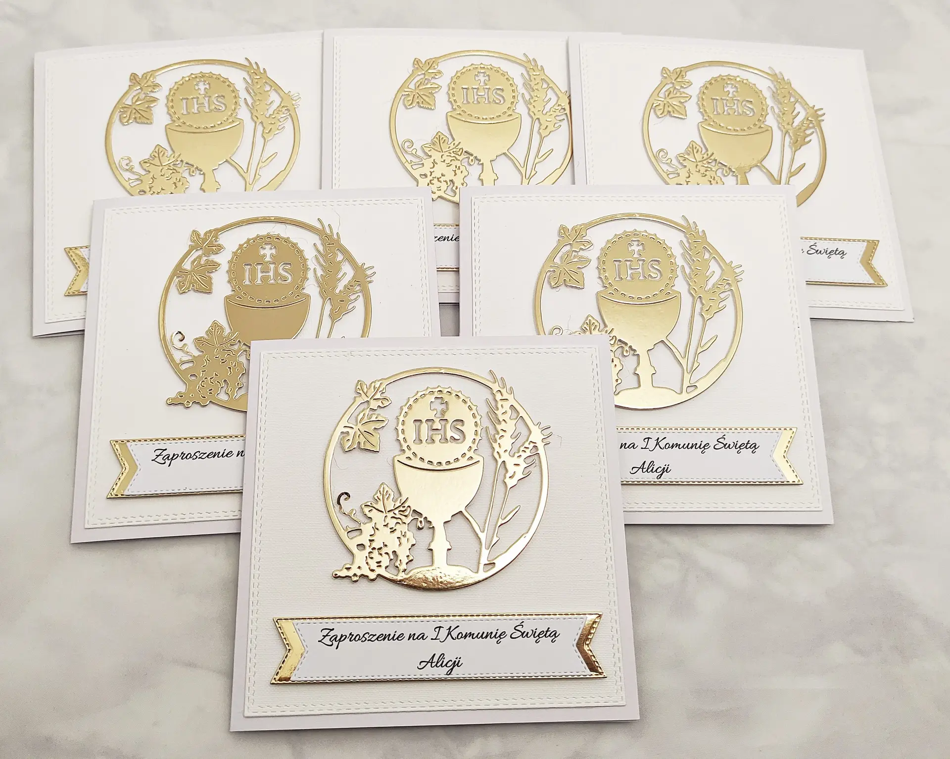 Kilka zaproszeń na Pierwszą Komunię Świętą z kielichem i symbolem IHS, bez złotego tła, ułożonych obok siebie, widok z góry, spójna seria biało-złotych zaproszeń.