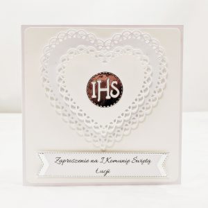 Biało-srebrne zaproszenie na Pierwszą Komunię Świętą z podwójnym ażurowym sercem i symbolem IHS, pojedyncze zaproszenie sfotografowane od frontu.