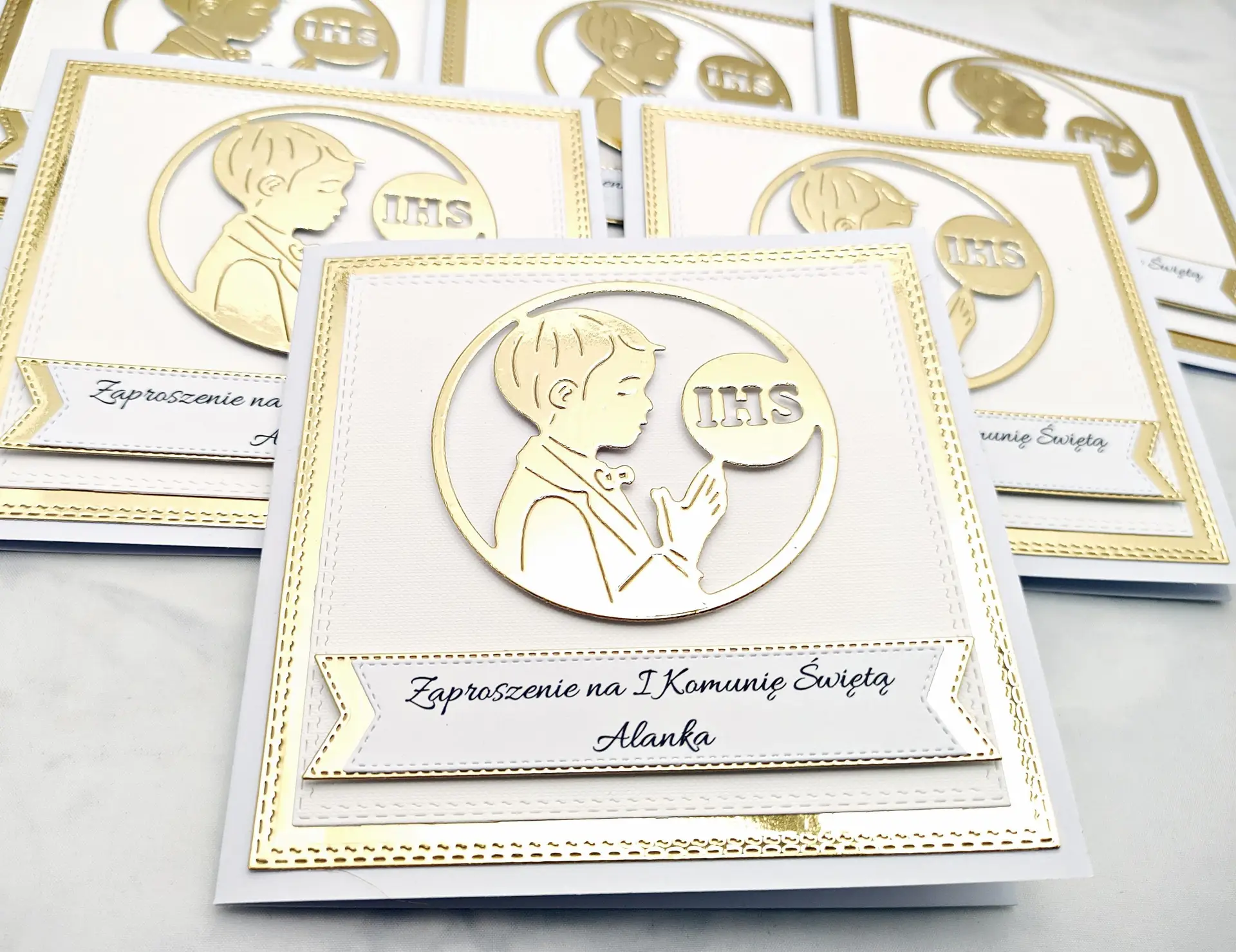 Kilka biało-złotych zaproszeń komunijnych dla chłopca z sylwetką chłopca i IHS, ułożonych obok siebie, widok z góry, lustrzane złote tło.