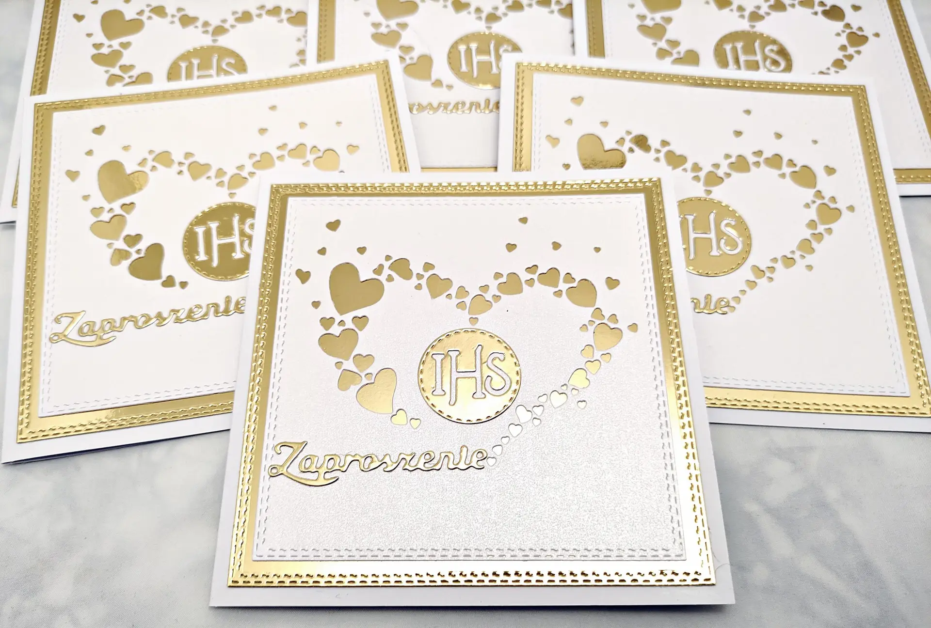 Kilka zaproszeń komunijnych z motywem serca z serduszek i symbolem IHS, ułożonych obok siebie, widok z góry w bliższym kadrze.
