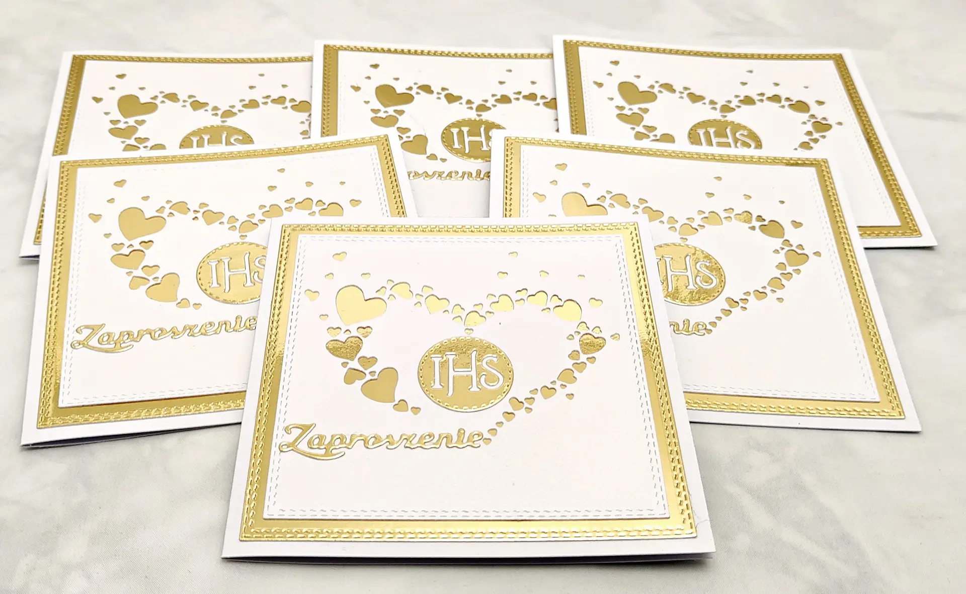 Zaproszenia na Pierwszą Komunię Świętą z sercem z serduszek i IHS, sfotografowane z góry z większej odległości, pokazujące kilka sztuk ułożonych obok siebie.