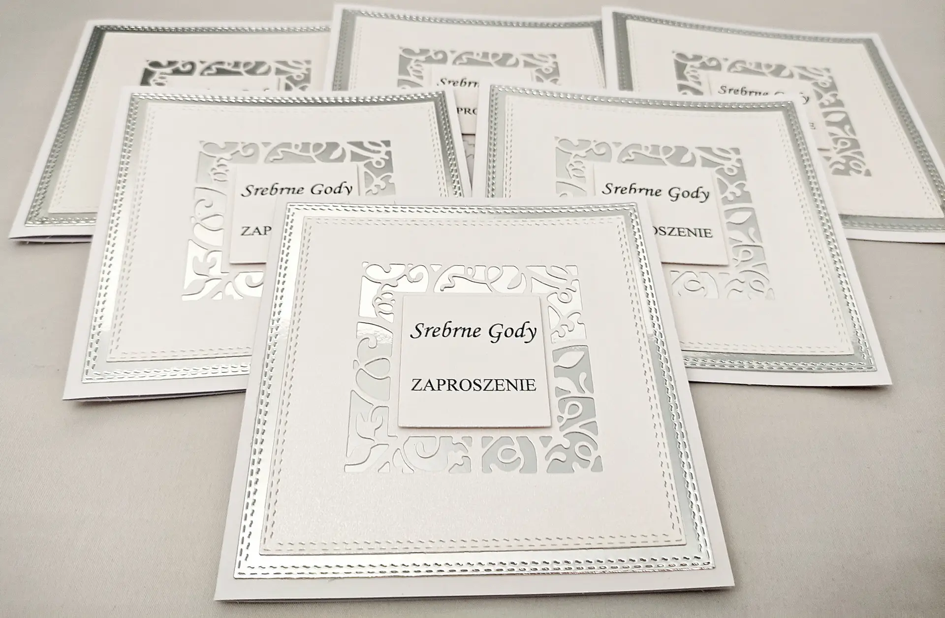 Zestaw zaproszeń na Srebrne Gody ułożonych obok siebie, na lustrzanym srebrnym tle, z kwadratową ażurową ramką i napisem „Srebrne Gody – Zaproszenie” – widok z góry