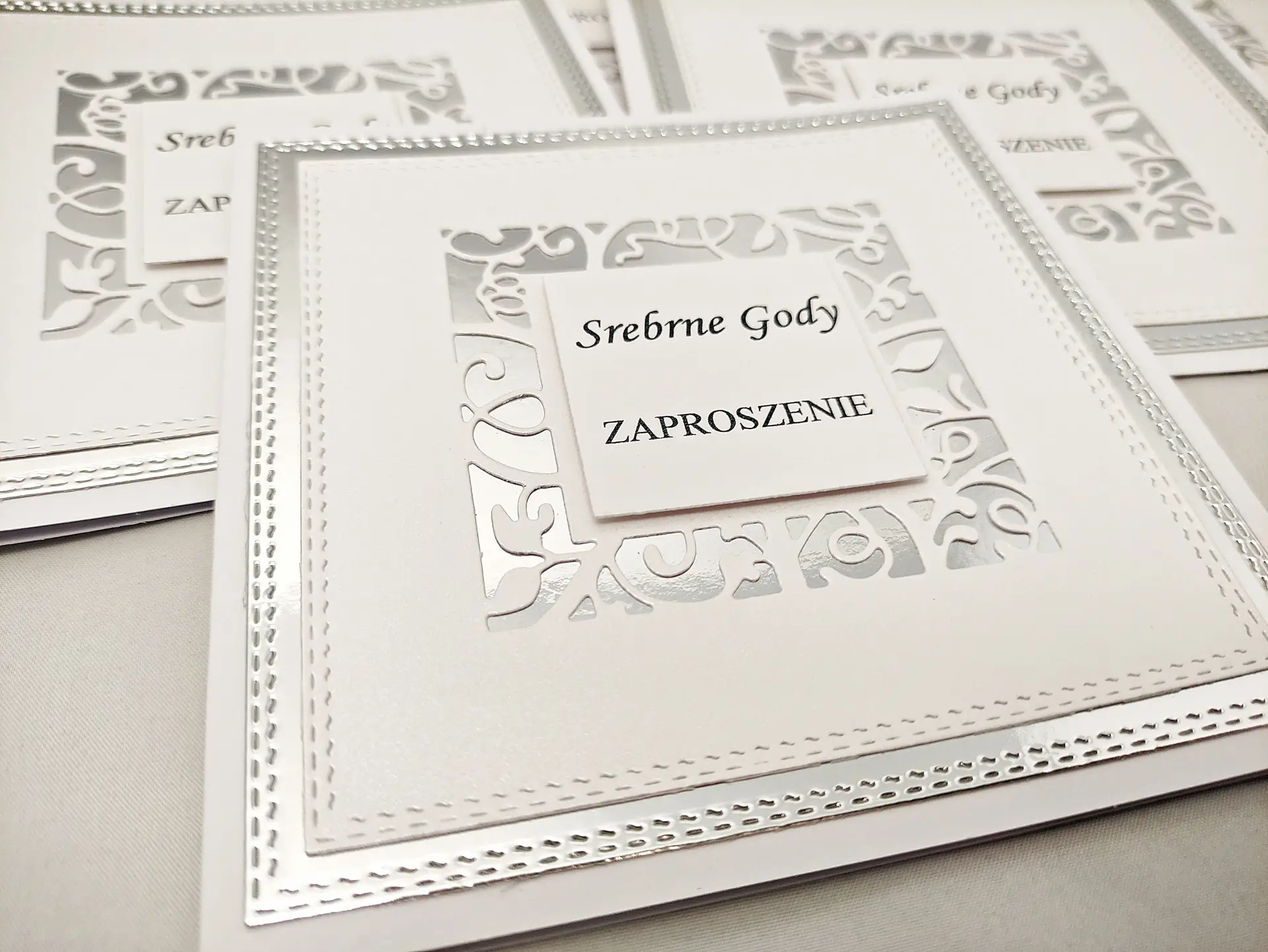 Zbliżenie detali zaproszenia na Srebrne Gody – ażurowa kwadratowa ramka na lustrzanym srebrze i elegancki nadruk „Srebrne Gody – Zaproszenie” na białym perłowym papierze
