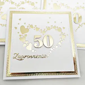 Eleganckie zaproszenie na 50 rocznicę ślubu na złotym tle, z białym perłowym papierem i sercem z małych serduszek wokół liczby 50 – widok z góry